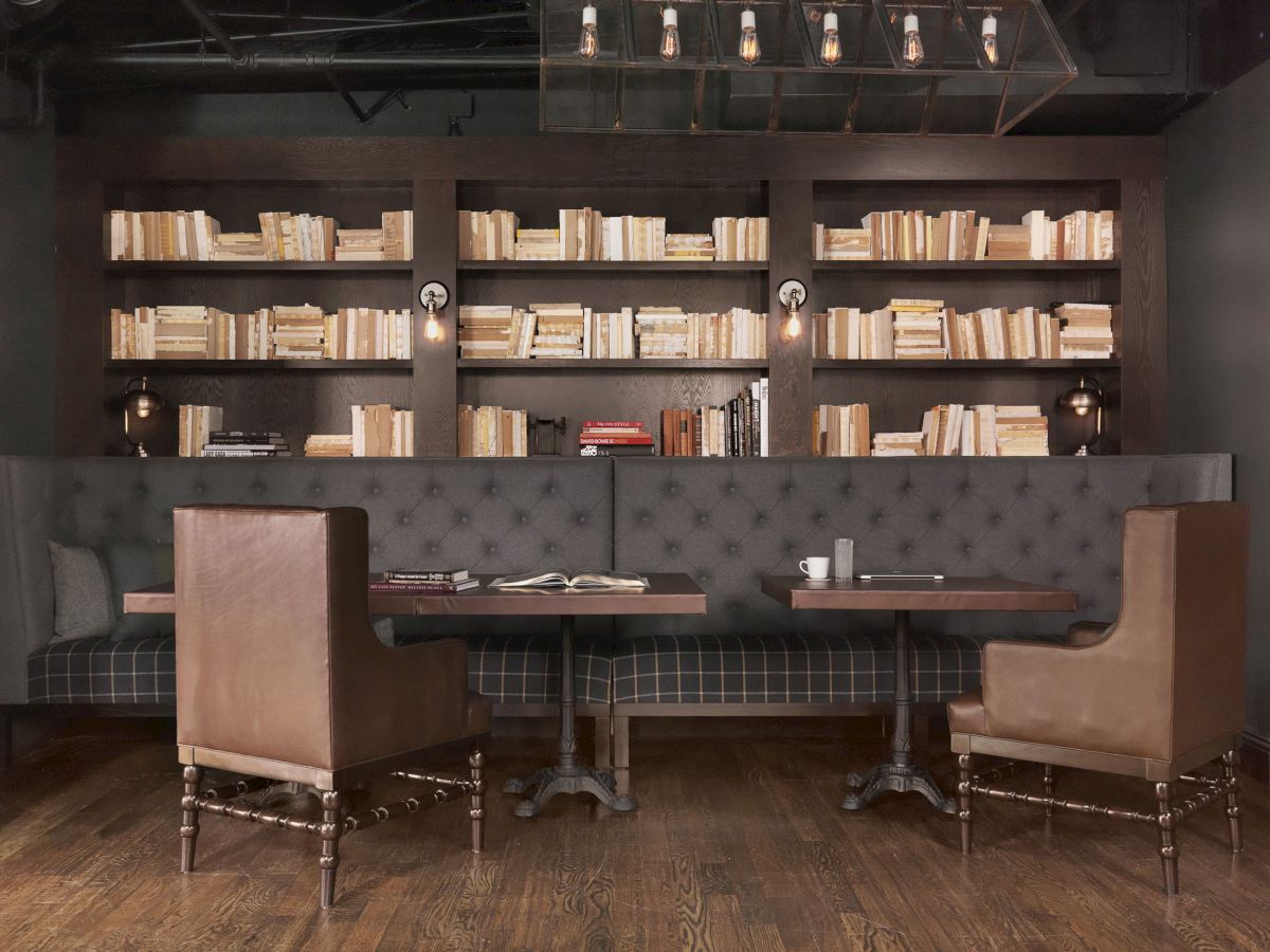 A cozy library-themed cafe with a dark, tufted sofa, wooden tables, leather chairs, and bookshelves filled with books on the wall.