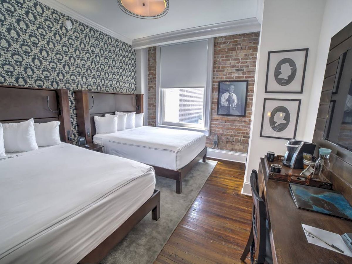 The image shows a hotel room with two beds, a desk with a lamp and chair, patterned wallpaper, and framed art on a brick wall.