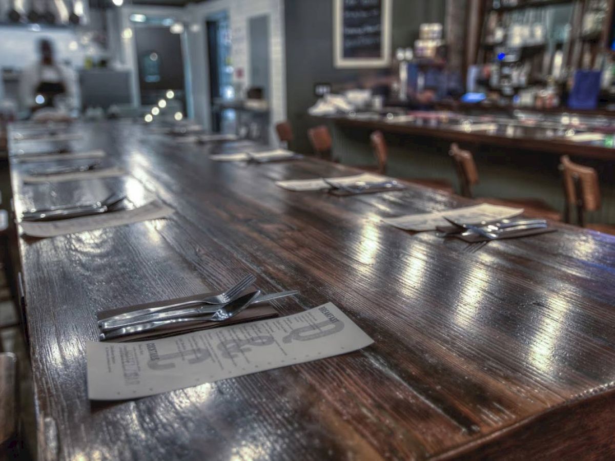 A long wooden table is set with placemats, forks, spoons, and knives, while a bar area in the background features stools and illuminated bottles.