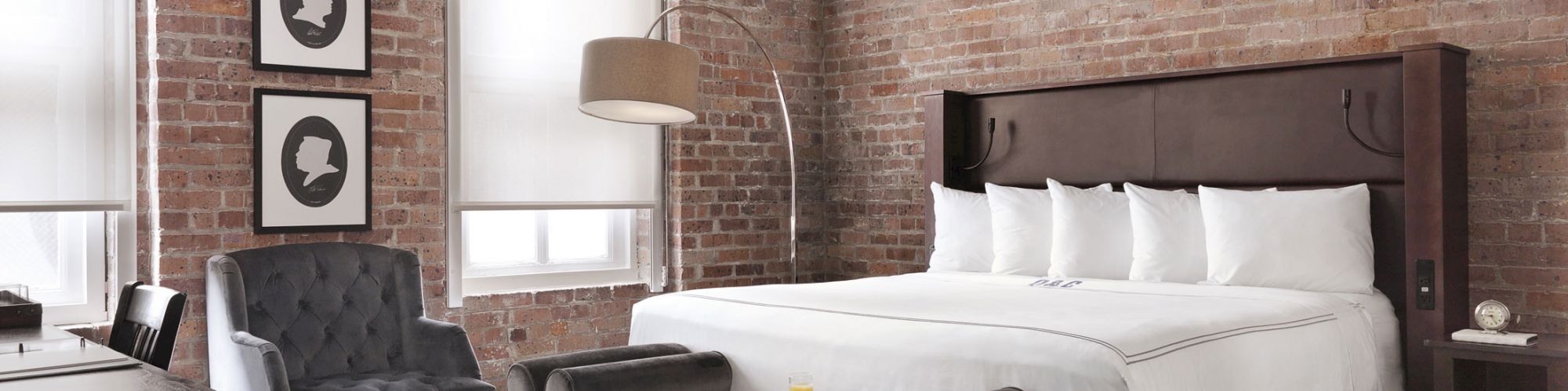 A modern bedroom with a brick wall, king-sized bed, two gray chairs, a stylish light fixture, and a patterned carpet, featuring minimalist decor.