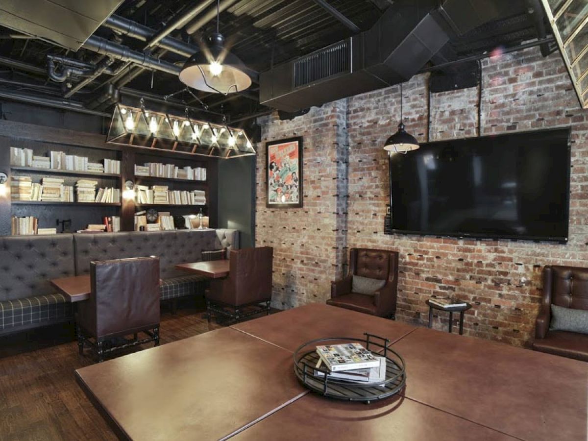 A cozy, industrial-style room with exposed brick walls, a large TV, leather chairs, a bookshelf, and a central table with magazines.