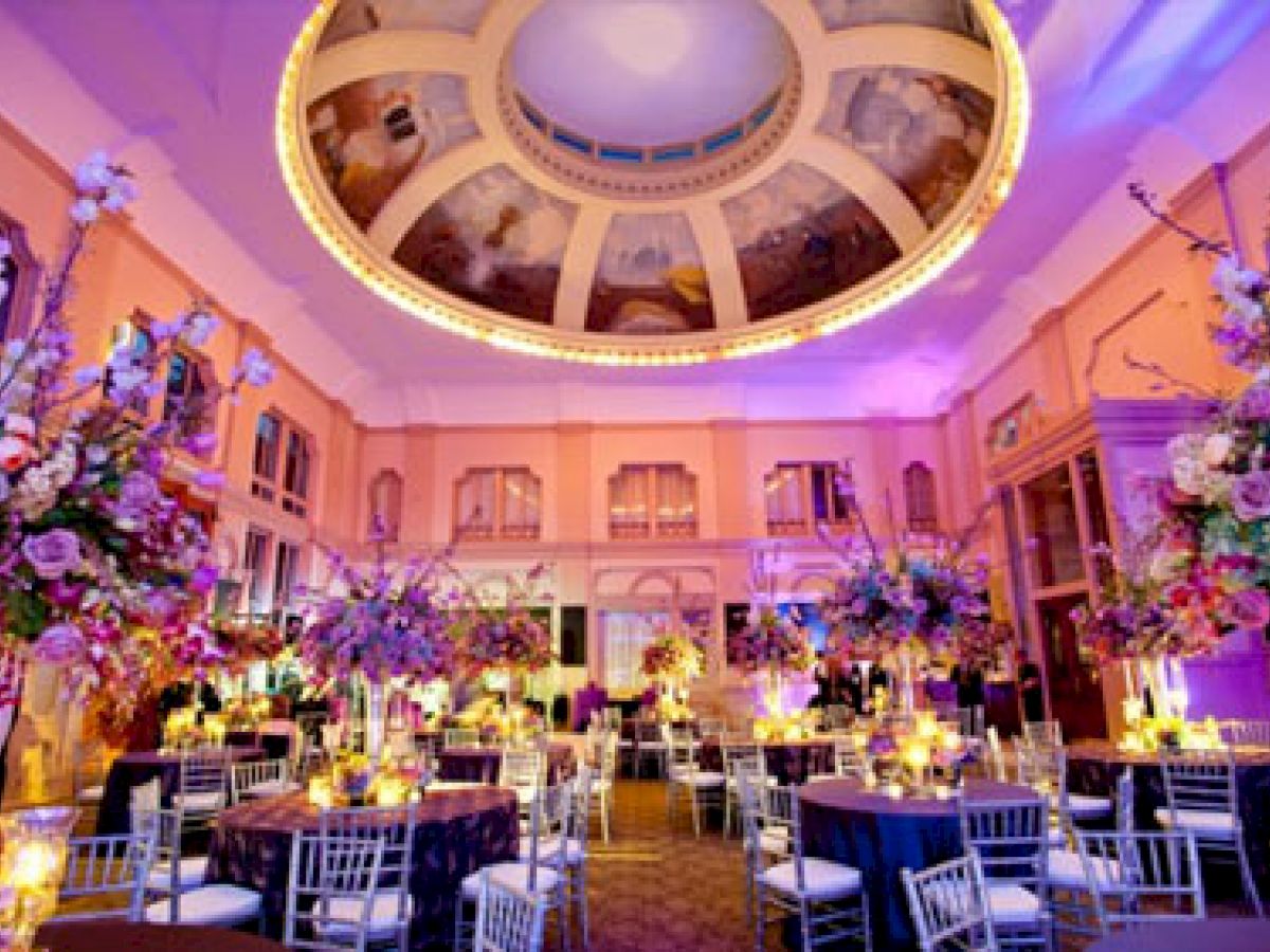 A lavishly decorated ballroom with grand floral arrangements, elegant table settings, and a stunning ceiling with intricate artwork and lighting.