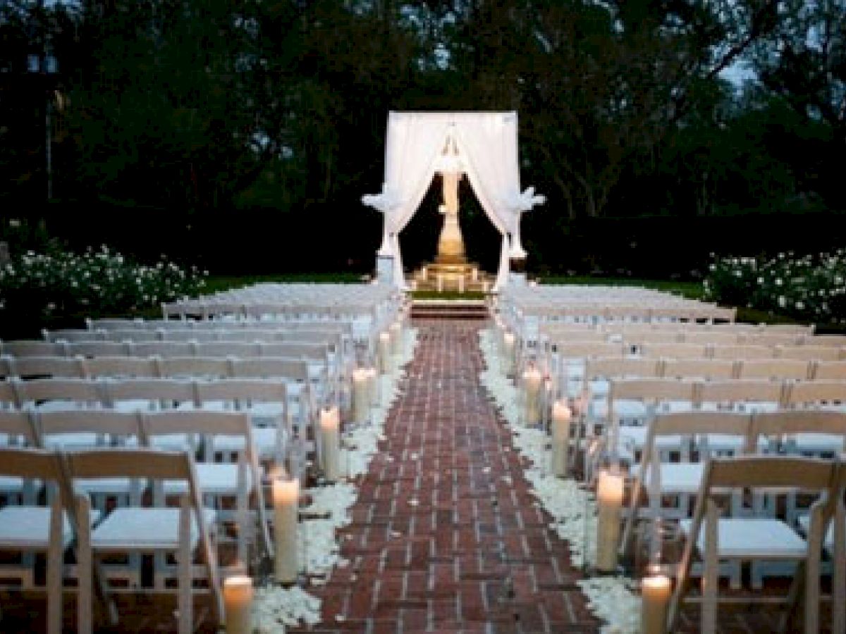 An outdoor wedding setup with white chairs aligned along a brick aisle, decorated with petals and candles leading to a draped altar at the front.