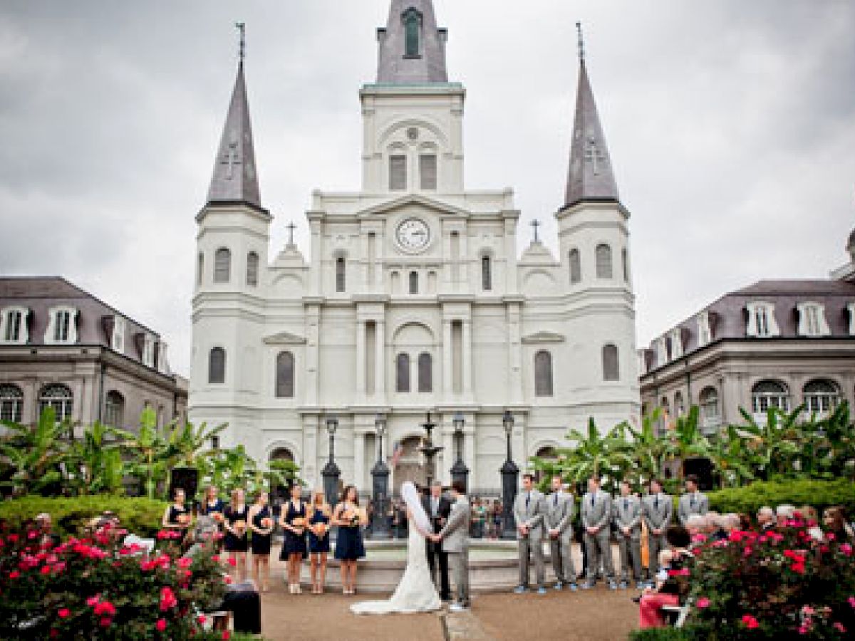 A wedding ceremony is taking place in front of a large, ornate white church with two steeples and prominent clock, surrounded by greenery and flowers.