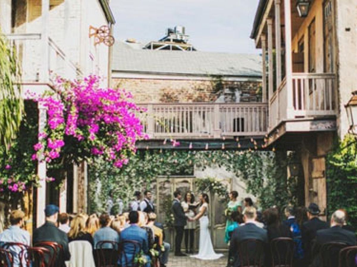 A wedding ceremony is taking place in an outdoor courtyard with guests. The space is adorned with greenery and bright flowers, and there are balconies.