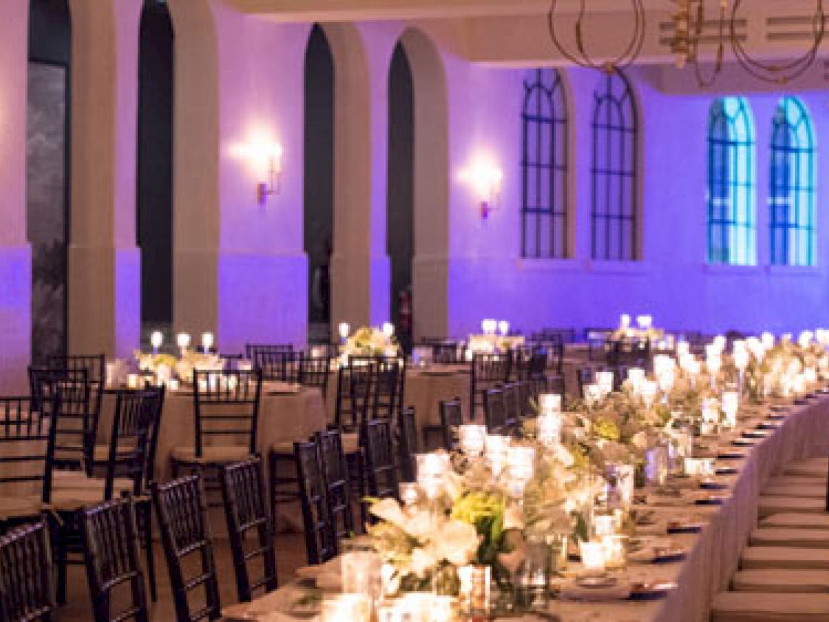 The image shows an elegant reception hall set for an event, with long tables adorned with candles and flowers, and rows of chairs.