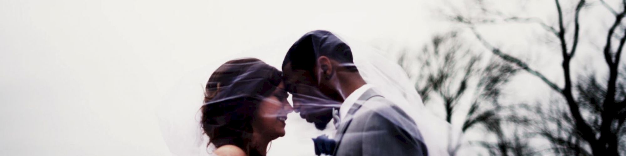 A bride and groom stand closely, forehead to forehead, with a veil draped over them. Trees in the background are bare, suggesting a winter setting.