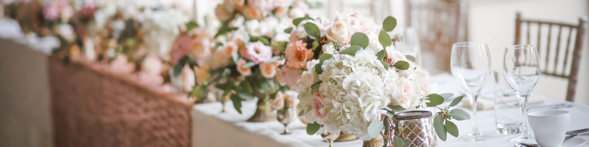 The image shows a beautifully decorated table with floral arrangements, wine glasses, a teacup, and elegant tableware, likely set for a formal event.