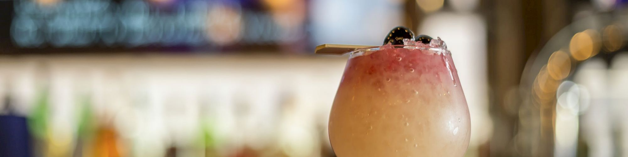 A cocktail glass with a layered red and white drink garnished with cherries on a bar counter, with a blurred figure on TV in the background.