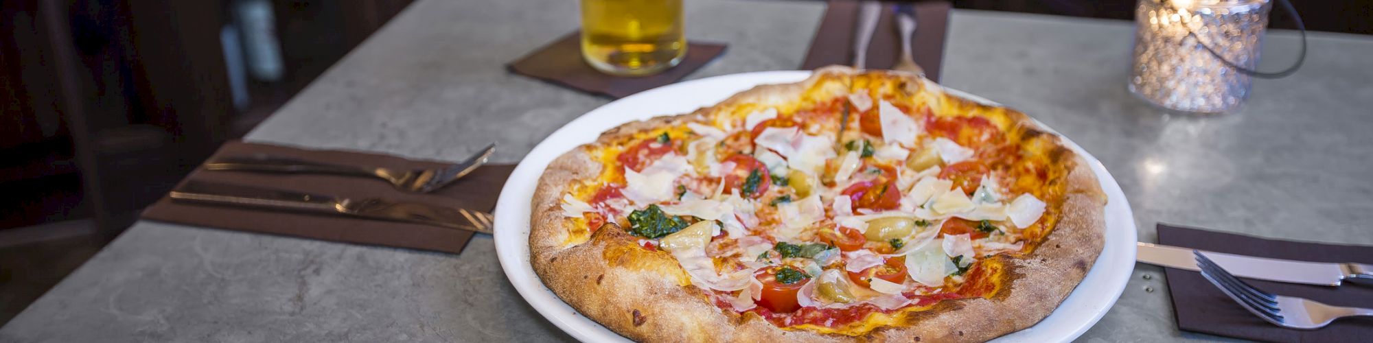 A delicious pizza on a plate, set on a table with utensils, a beer, and a lit candle in the background.