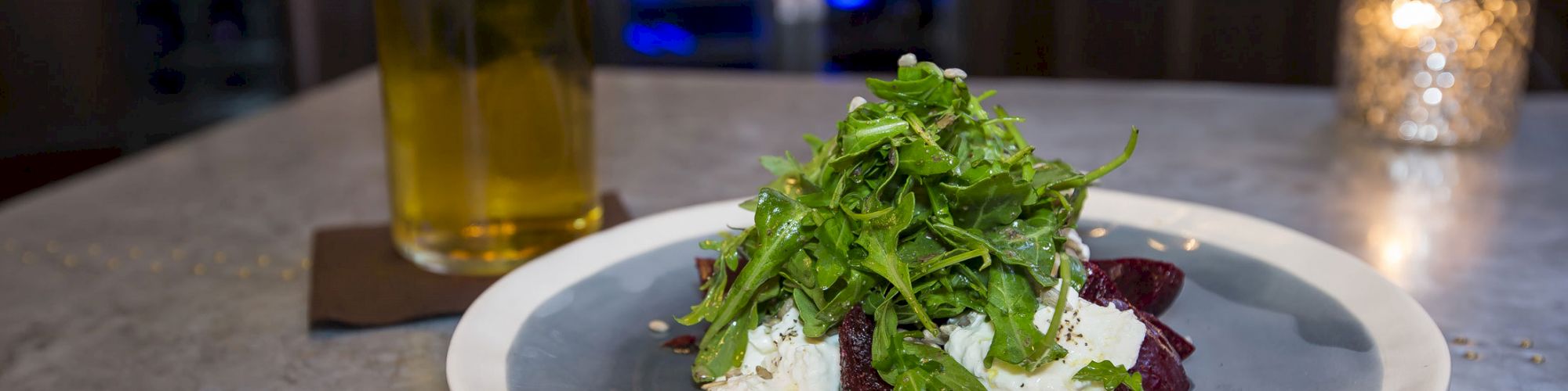 A plate of food with greens, possibly arugula, and another topping, sits next to a glass of beer on a table with a lit candle in the background.