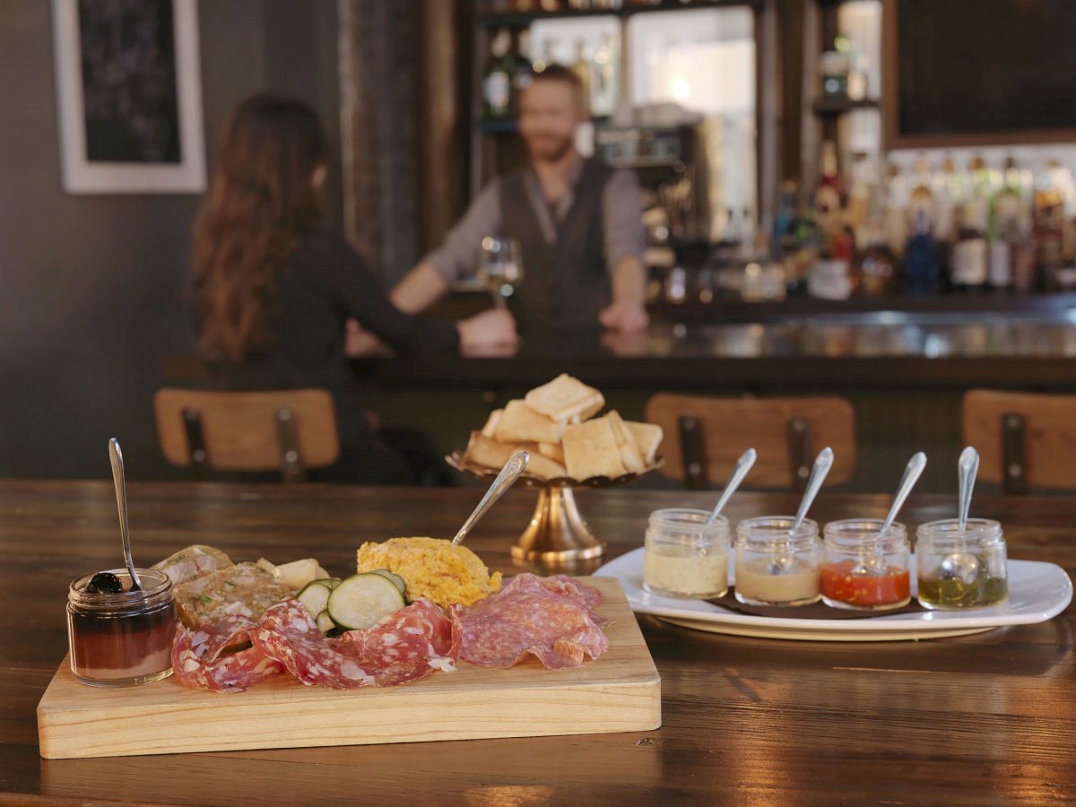 A charcuterie board, bread, and various sauces are on a bar counter, with two people talking in the background.