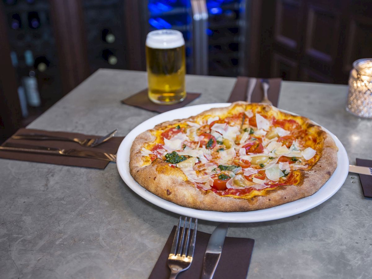 A pizza topped with vegetables on a restaurant table alongside a pint of beer, a candle, cutlery, and napkins, with a wine bottle rack in the background.