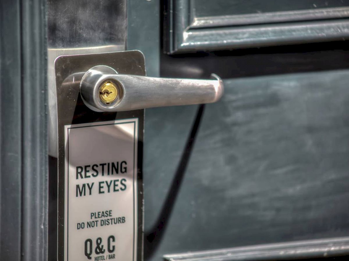 A door handle with a sign that reads "Resting My Eyes. Please Do Not Disturb. Q & C Hotel Bar." The handle is silver, and the door is dark.