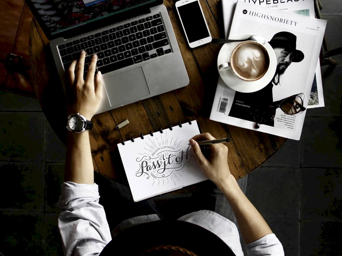 A person works at a wooden table with a laptop, phone, magazines, headphones, a cup of coffee, and a notebook displaying handwritten text.