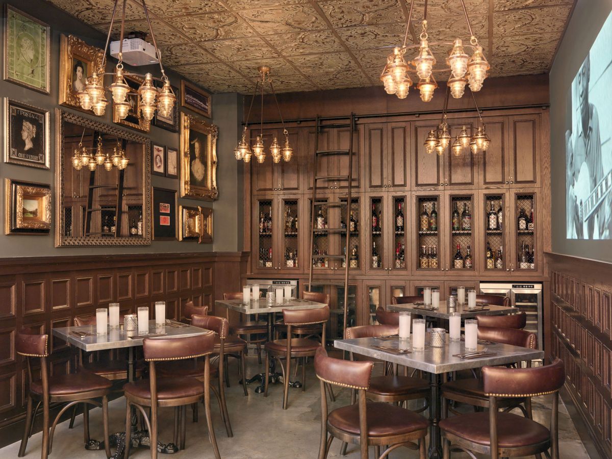 A cozy room with wooden paneling, vintage chandeliers, framed artworks, shelves of books and bottles, and tables with candles.