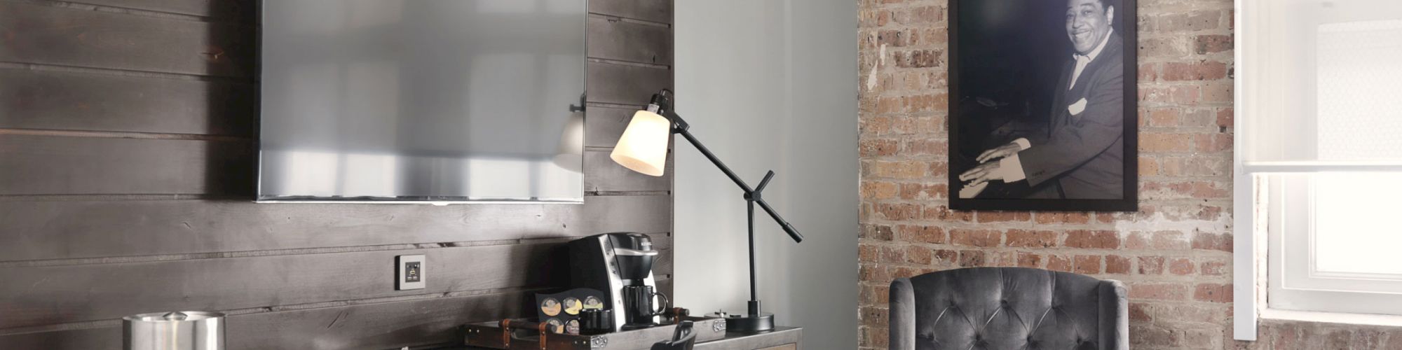 A modern hotel room features two beds, a wall-mounted TV, a desk with a lamp, a chair, and framed artwork on an exposed brick wall.