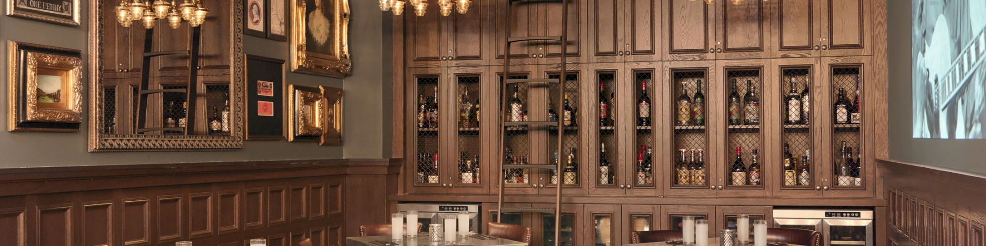 A vintage-style restaurant interior with wooden wall panels, framed pictures, chandeliers, and candle-lit tables with leather chairs.