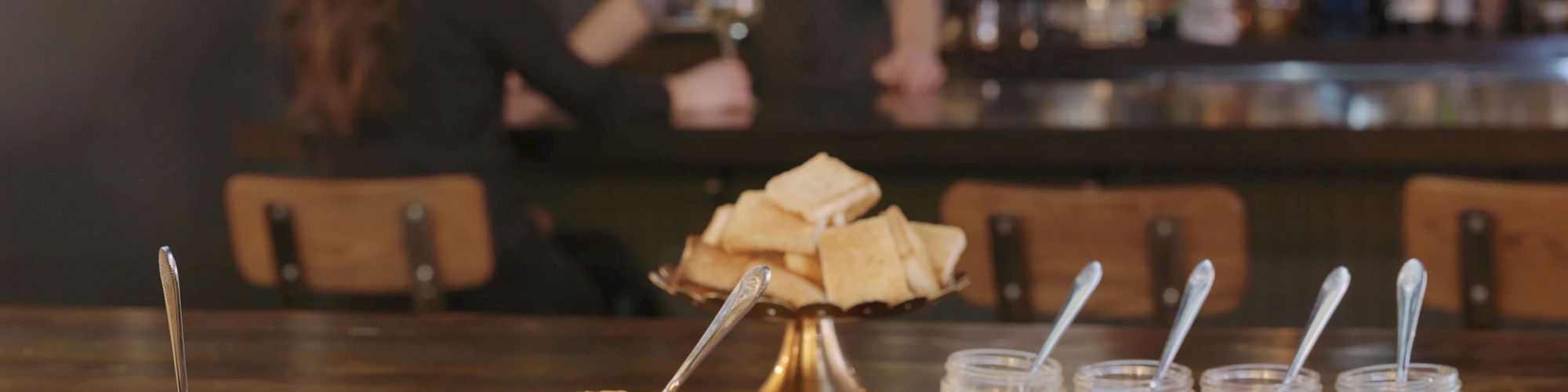 A charcuterie board with meats, jars of condiments, and breadsticks is on a bar table; two people are conversing in the background.