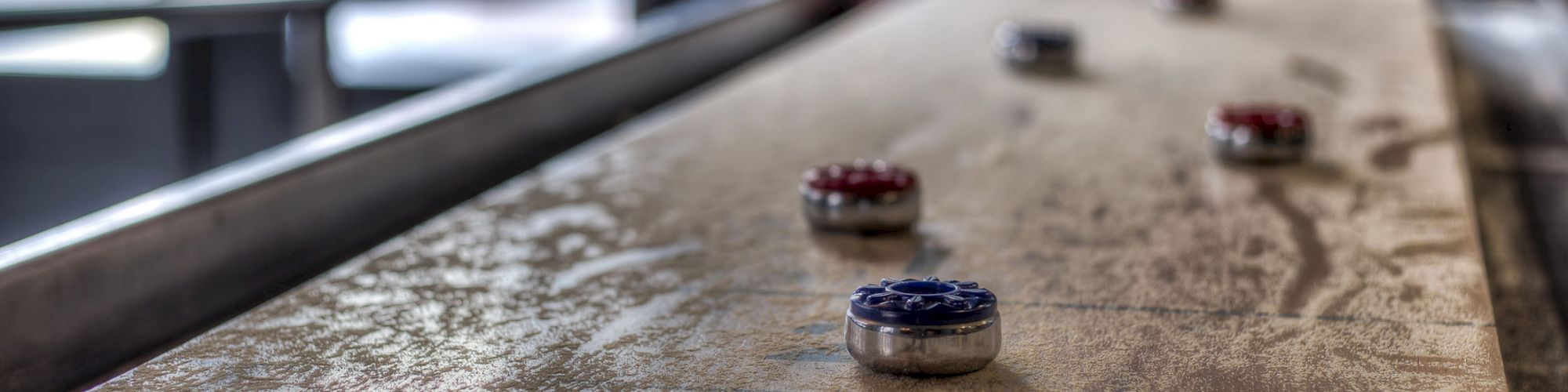 A close-up of a shuffleboard table with red and blue pucks placed on it, seen in an indoor setting with blurred background details.