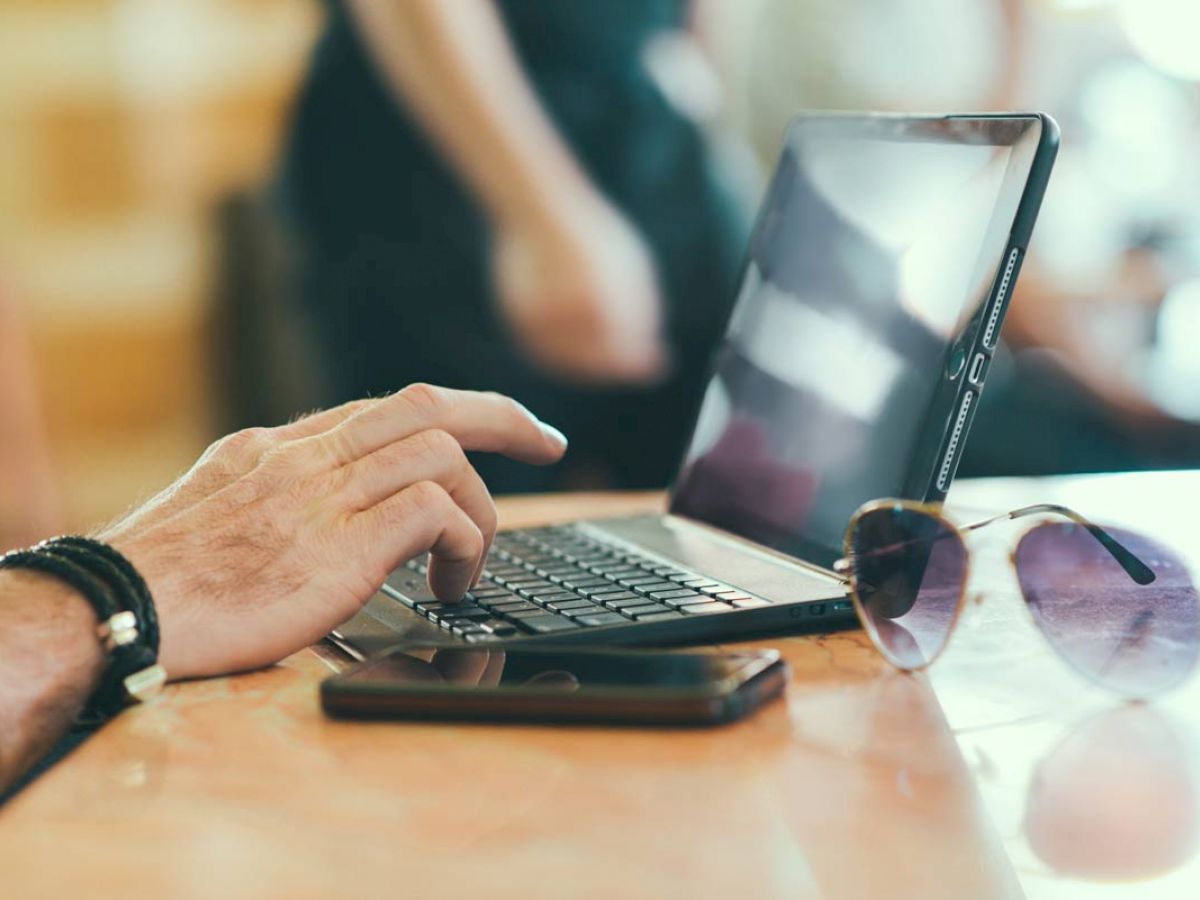 A person using a laptop, with a smartphone and sunglasses resting on the table beside them.