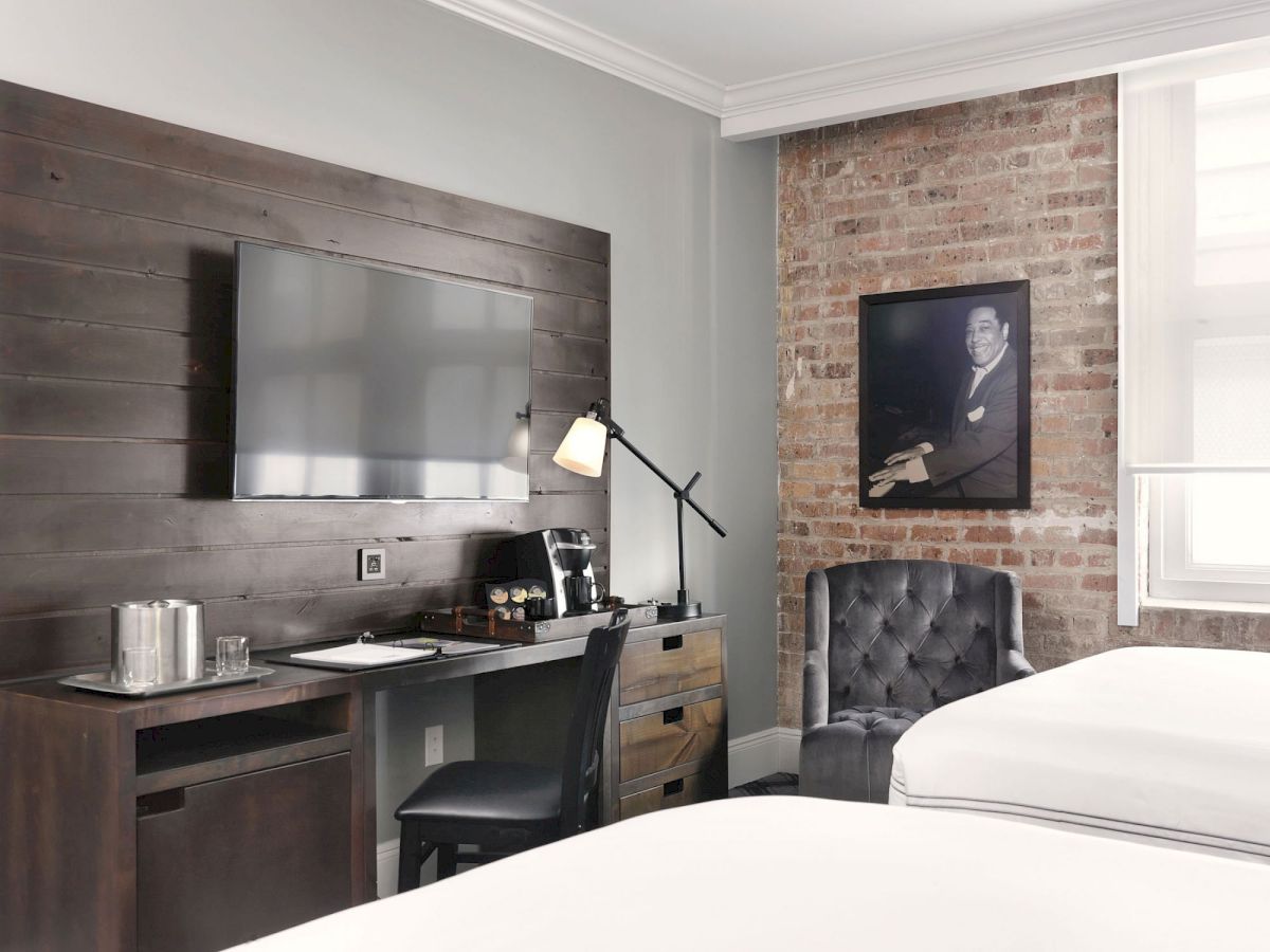 A small modern kitchenette with a sink, coffee maker, and desk lamp, plus a brick wall with a framed portrait and a chair.