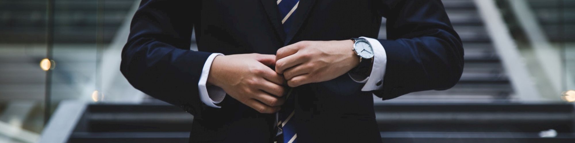 A person in a suit stands in front of modern staircase, adjusting their jacket, with a watch on their wrist.