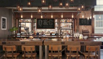 A modern bar setup with wooden tables, chairs, and hanging lights. Bottles and glassware are displayed behind the bar with a "Q&C Hotel/Bar" sign ending the sentence.