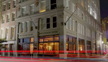 A multi-story building on a street corner, with light trails from passing vehicles and illuminated windows showing interior details.