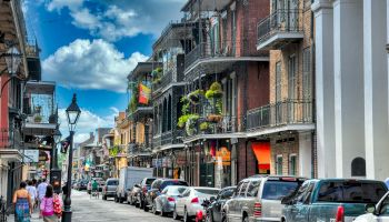 A colorful street scene with parked cars, historic buildings with balconies, hanging plants, and people walking on the sidewalk under a blue sky.