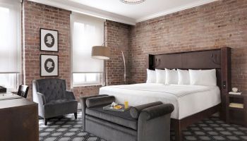 This image shows a stylish bedroom with a brick wall, king-size bed, a cozy armchair, and a small sofa with a tray of breakfast on it, in a hotel room.