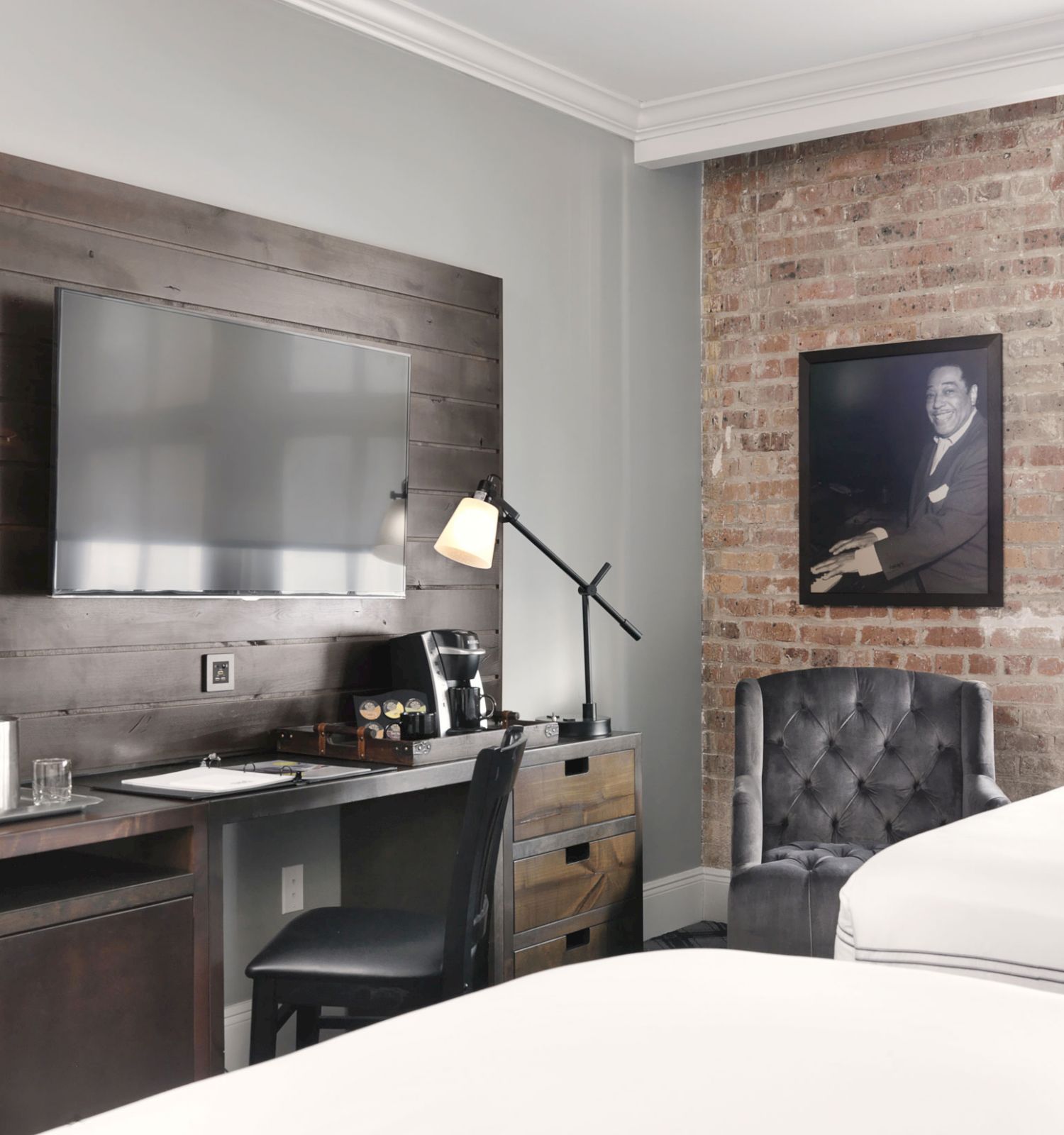 A modern hotel room features a wall-mounted TV, desk with a lamp and coffee maker, a tufted chair, exposed brick wall, and a photo.