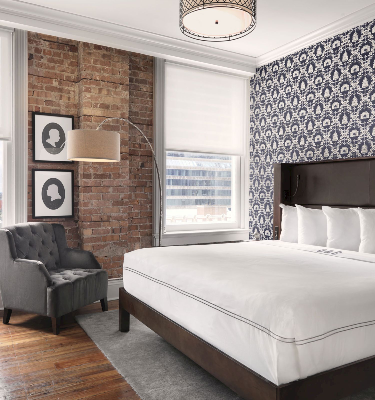 Modern bedroom with patterned wallpaper, a brick accent wall, and large windows. There’s a king-sized bed, armchair, desk, and wall art.