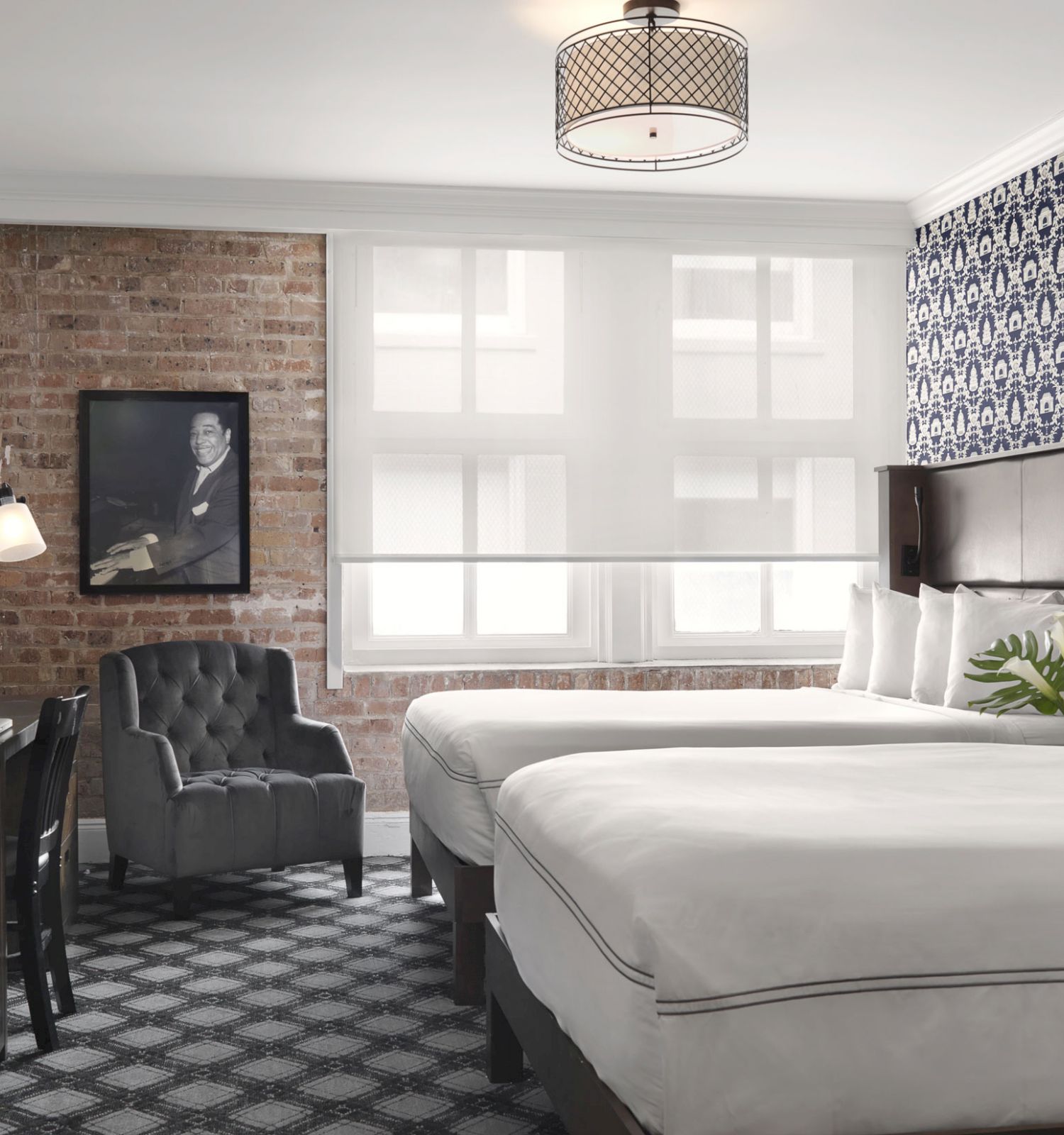 A stylish hotel room features two beds, brick and patterned walls, a gray armchair, a desk, modern lighting, and a framed black-and-white photo.