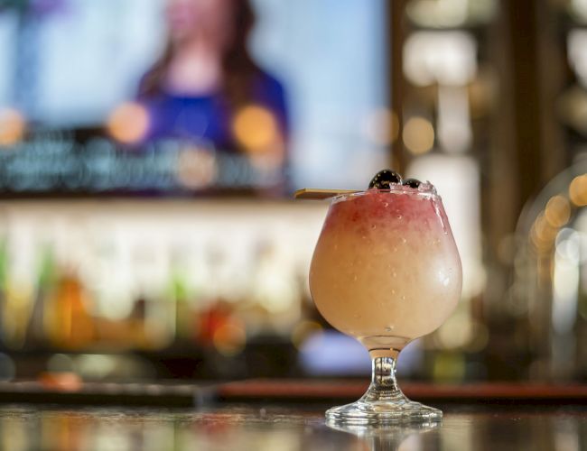 A cocktail in a frosty glass garnished with a cherry sits on a bar counter with a blurry TV screen and bar shelves in the background.