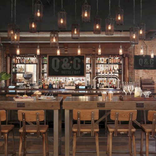 The image shows a modern bar area with wooden tables, chairs, hanging light fixtures, and bottles displayed on the shelves.