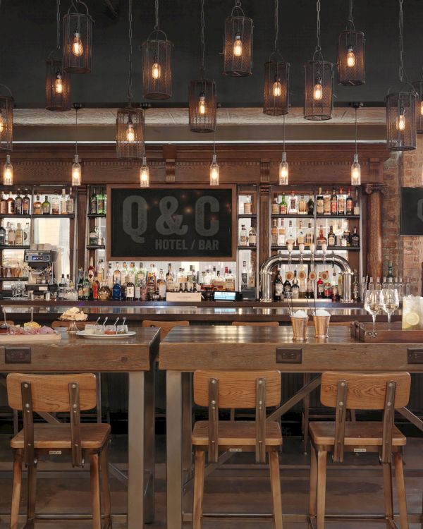 The image shows a modern bar area with wooden tables, chairs, hanging light fixtures, and bottles displayed on the shelves.