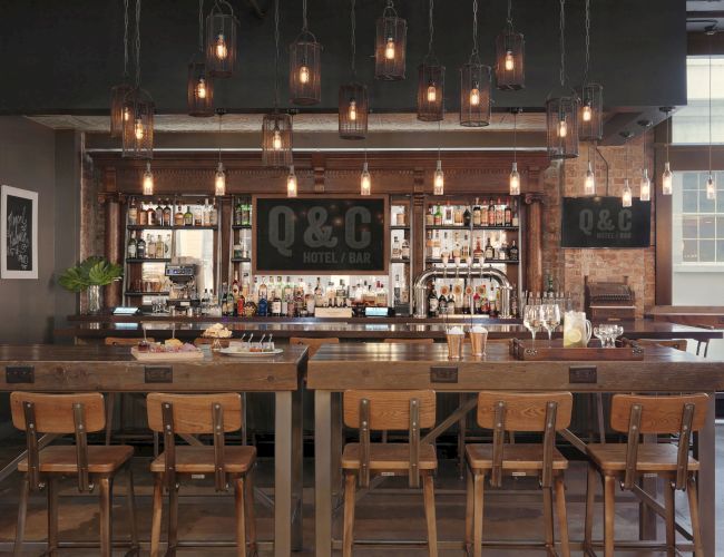 The image shows a modern bar area with wooden tables, chairs, hanging light fixtures, and bottles displayed on the shelves.