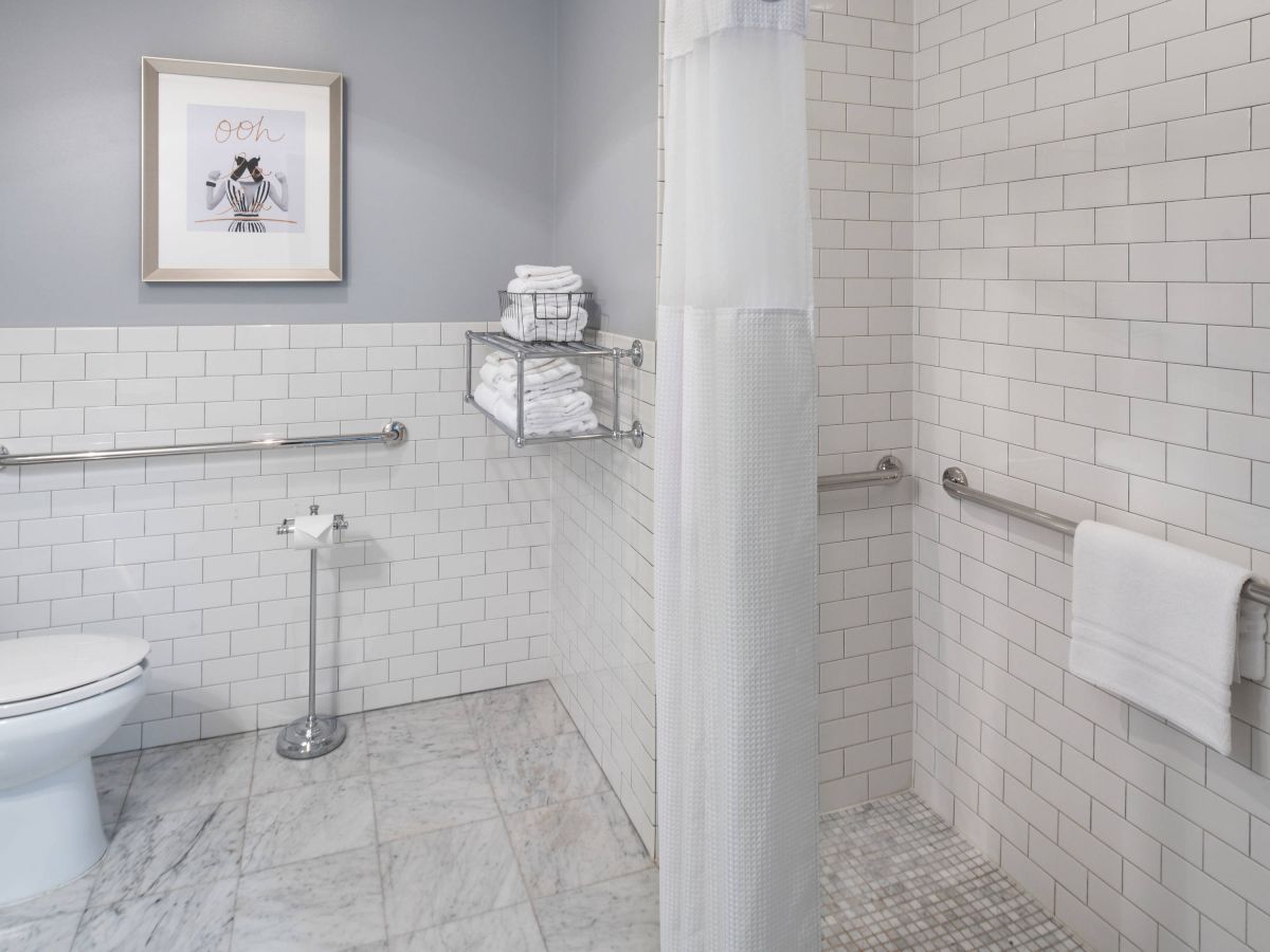 This image shows a clean, modern bathroom with white tiles, a toilet, a shower with a curtain, grab bars, and neatly folded towels on shelves.