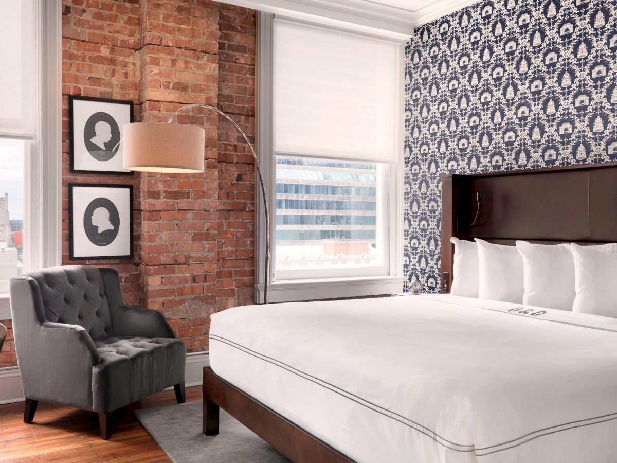 A stylish hotel room featuring a king-sized bed, accent chair, brick wall, framed silhouettes, and a blue patterned accent wall.