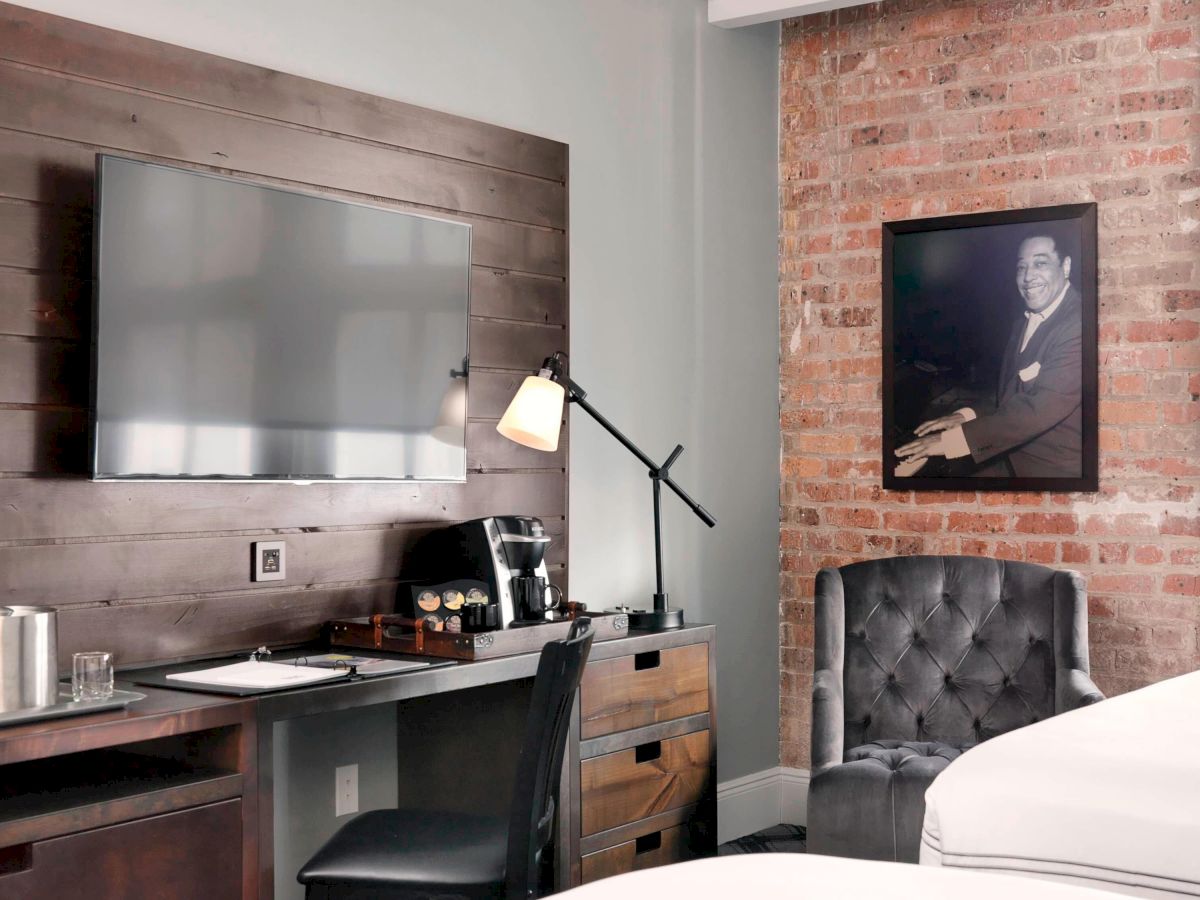 A cozy bedroom features a wall-mounted TV, desk with lamp, coffee maker, and cushioned chair near an exposed brick wall with a framed portrait.