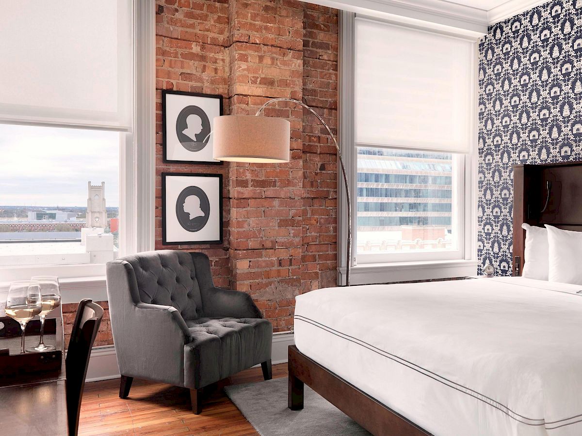 The image shows a stylish hotel room with a king-size bed, patterned wallpaper, brick accent wall, gray chair, framed silhouette art, and large windows.