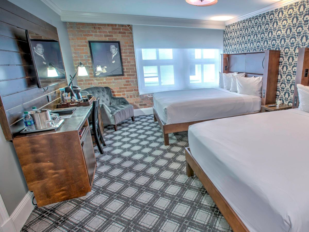 The image shows a hotel room with two large beds, a wooden dresser, a patterned accent wall, a window, and framed pictures on the walls.