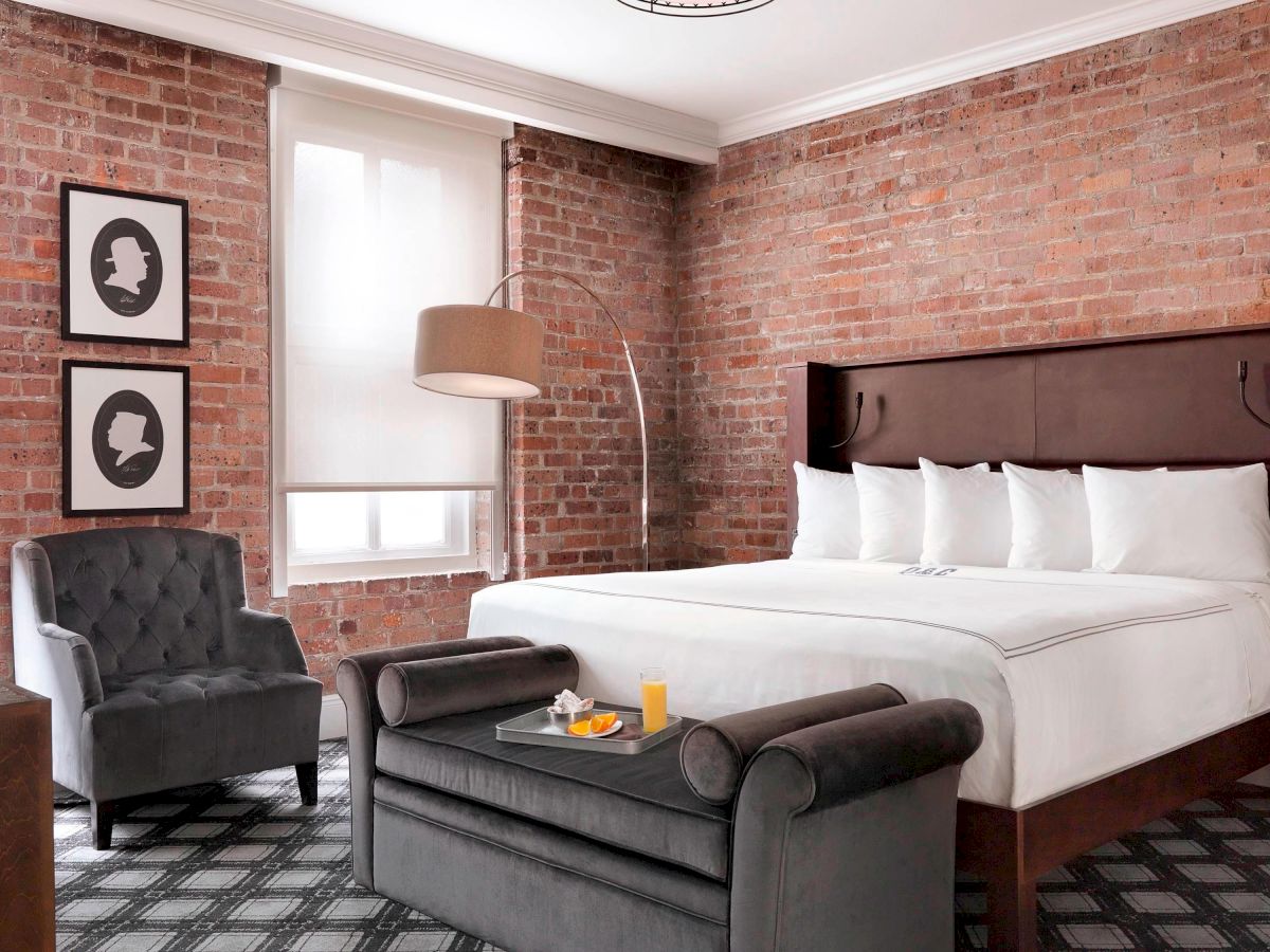 A stylish bedroom with an exposed brick wall, a large bed, a gray armchair, a table with food, and modern decor, including silhouette art on the wall.