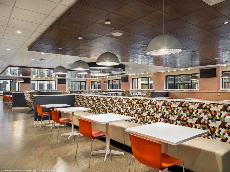 A bright cafeteria with long white tables, orange chairs, colorful tiled banquette, pendant lights, and a calm, spacious dining area.