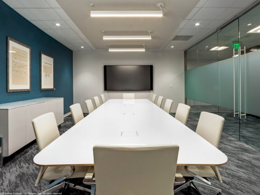 A modern conference room with a long white table, white chairs, a wall-mounted screen, blue accent wall, and glass doors in a sleek office setting.