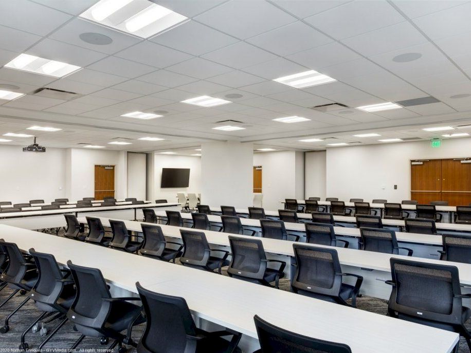 A large, empty conference or lecture room with rows of black chairs and long white desks, bright ceiling lights, and a few doors at the back.