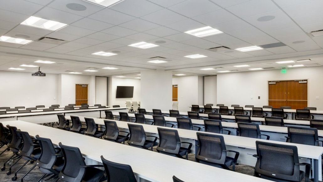 A large, empty conference room with long white desks, black chairs, and ceiling lights; it appears ready for a lecture or meeting.