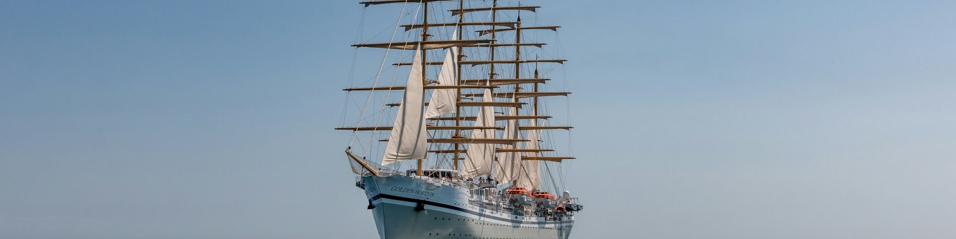 A tall schooner with multiple masts sails calmly on a calm sea, reflecting in the water.