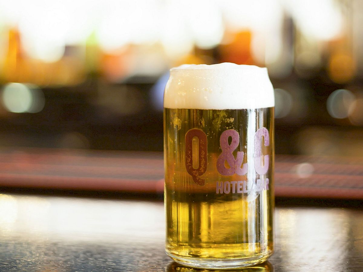 A frothy pint of beer sits on a table, labeled &ldquo;03 HOTEL BAR,&rdquo; with a blurred bartender and colorful lights in the background.